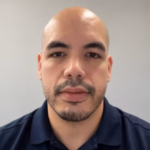 Close-up portrait of a Carlos with light stubble wearing a navy polo shirt, facing the camera with a calm, neutral expression.